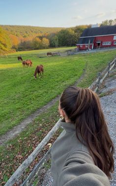 a woman standing in front of a fence looking at horses grazing on the field behind her