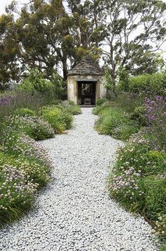 a gravel path leading to a small building surrounded by trees and bushes with purple flowers in the foreground