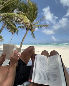 a man reading a book on the beach under a palm tree while laying in a hammock