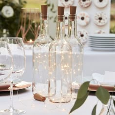 three wine bottles with lights in them sitting on a table next to plates and glasses