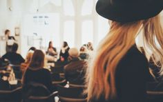 a woman with long blonde hair standing in front of a room full of other people