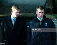 two men standing next to each other in front of a brick wall wearing coats and ties