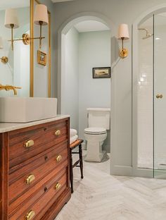 a bathroom with a walk in shower next to a white toilet and wooden dresser under a gold framed mirror