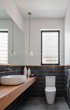 a white toilet sitting next to a bathroom sink under two windows in a room with black tiles on the walls