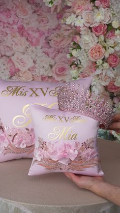 two pink pillows with gold lettering on them and flowers in the background, one has a tiara