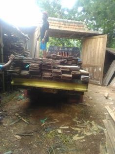 two men are standing on the back of a truck full of wood planks in front of a shed