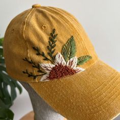 a yellow hat with embroidered flowers on the front and side, sitting next to a potted plant