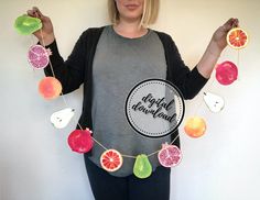 a woman standing in front of a white wall holding fruit garlands with the words happy holidays written on them