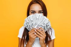 a woman covering her face with one hundred dollar bills in front of her face, on an orange background