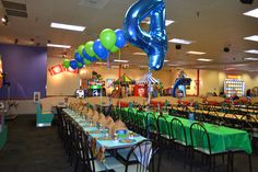 a room filled with tables covered in green and blue tablecloths, balloons and chairs