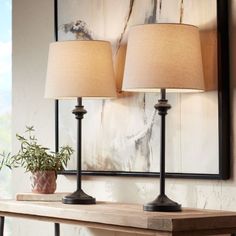 two lamps sitting on top of a wooden table next to a painting and potted plant