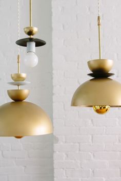 two lamps hanging from the ceiling in front of a white brick wall, one is gold and the other is black