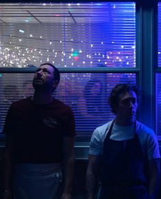 two men standing next to each other in front of a window with lights on it