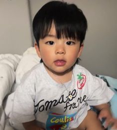 a little boy sitting on top of a bed wearing a white shirt and black hair