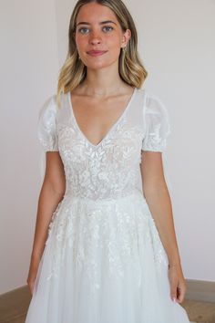 a woman wearing a white wedding dress standing in front of a wall and looking at the camera