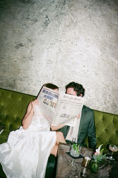 a man and woman sitting on a green couch reading the paper in front of them