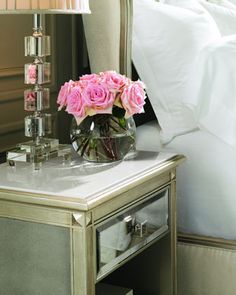 pink roses in a glass vase on a mirrored table next to a bed with white sheets