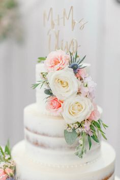 a white wedding cake with pink flowers and greenery on the top is decorated with gold lettering