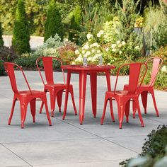 four red chairs around a small table in a courtyard area with flowers and bushes behind it