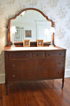 an old dresser with a mirror and two lamps on it in front of a floral wallpapered wall