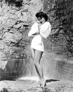 a woman in a short white dress standing next to a waterfall with her hands on her hips