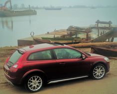 a red car parked next to a body of water with boats on the shore in the background