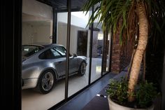 a silver porsche parked in front of a glass wall with palm tree and potted plant