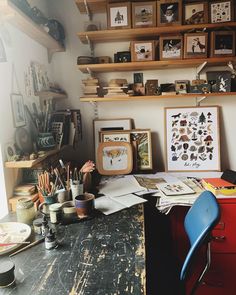 a messy desk with lots of art on the wall and shelves above it, along with other items