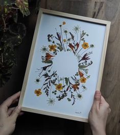 two hands holding up a framed print with flowers and leaves in the center on a wooden table