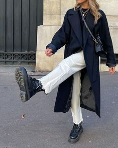 a woman in black coat and white pants walking down the street with her foot up