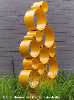 a tall yellow sculpture sitting in the grass next to a brick wall and green plants