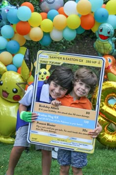 two young boys holding up a pokemon birthday party sign in front of balloons and streamers