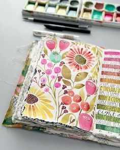 an open book with flowers on it next to some watercolor paints