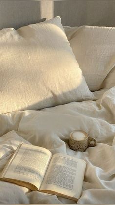 an open book is laying on a bed with white sheets and pillows, next to a coffee cup