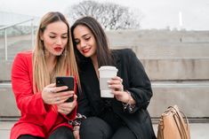 two young women sitting on steps looking at their cell phones while holding coffee cups in front of them