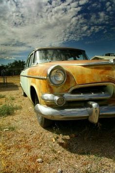 an old rusted car sitting in the middle of a dirt field under a cloudy blue sky