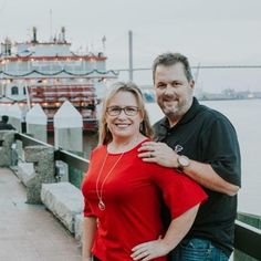 a man and woman standing next to each other on a pier near the water with a boat in the background