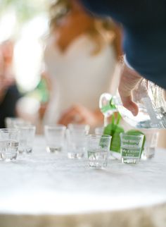 a person pouring water into shot glasses on a white tablecloth with people in the background