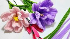 three different colored flowers sitting on top of a white table next to purple and green stems