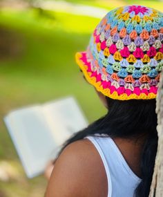 a woman wearing a colorful crocheted hat and reading a book while leaning against a tree