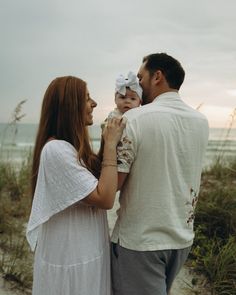 a man and woman holding a baby on the beach at sunset with sea oats in the background