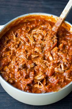 a white pot filled with chili and meat on top of a wooden table next to a spoon