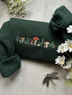 a green sweater with embroidered mushrooms and flowers on the side next to white daisies