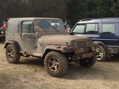 an old jeep is parked next to a blue truck in the dirt with trees in the background