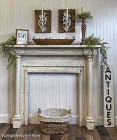 a white fireplace mantel with candles and pictures on the wall above it, along with a basket filled with greenery