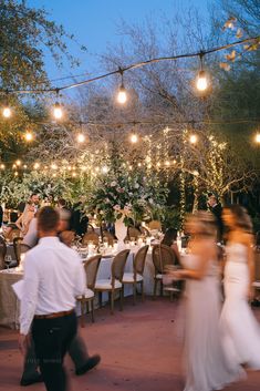 people are walking through an outdoor dining area at night with lights strung from the trees