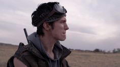 a man with goggles and a hat on standing in an open field, looking off into the distance