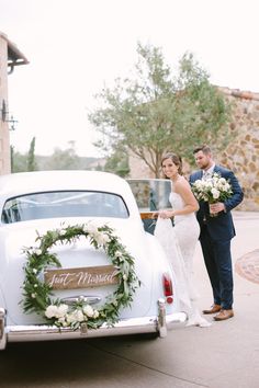 a bride and groom standing next to an old car with a wreath on the back