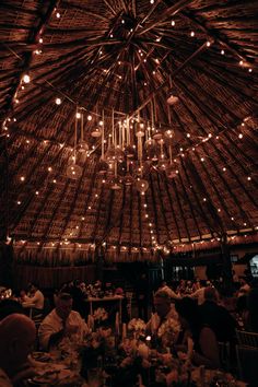 people are sitting at tables under a straw roof with lights hanging from the rafters