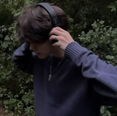 a young man wearing headphones while standing in front of some bushes and trees with his hands to his ears